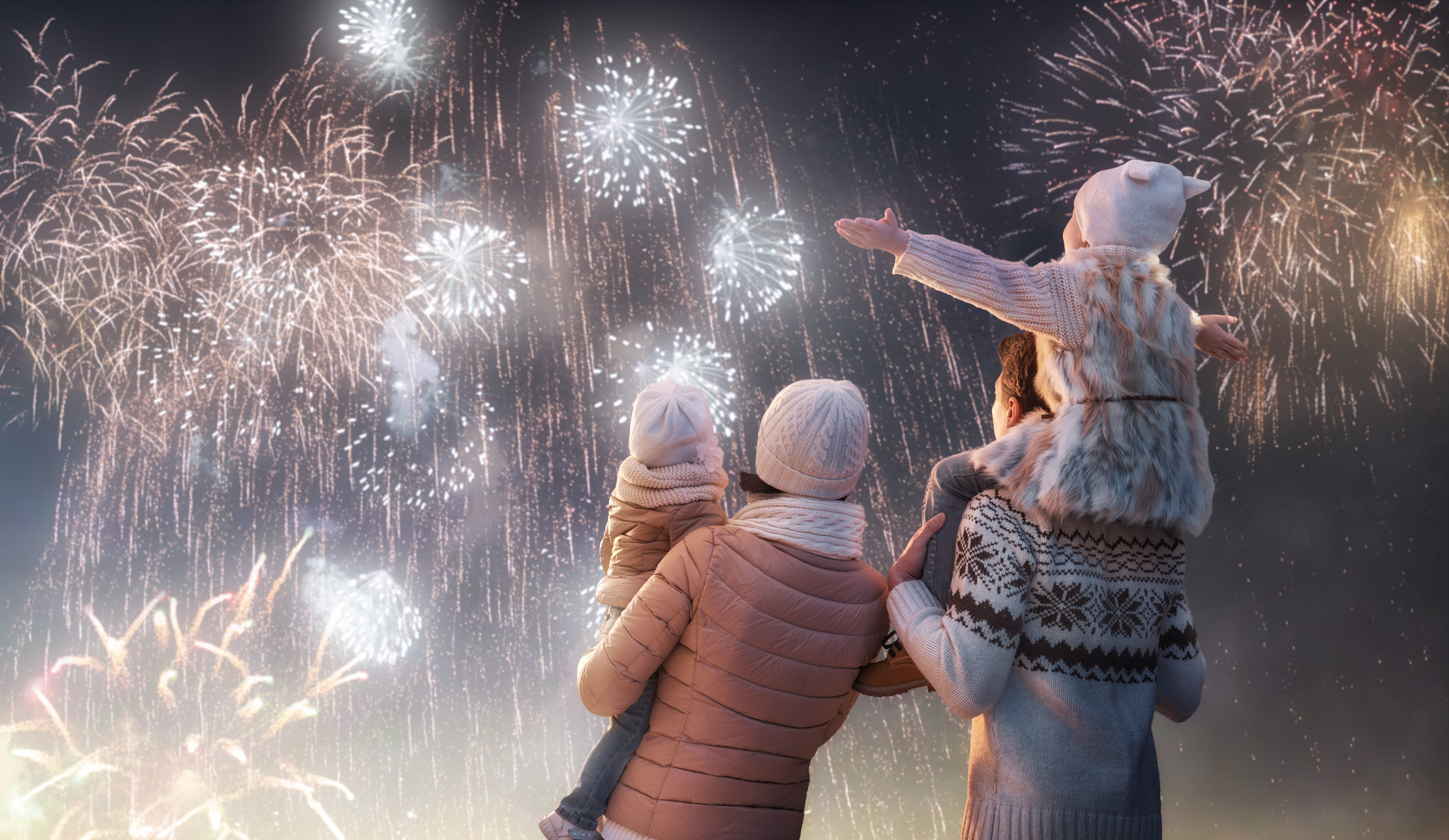 New Year holiday season. Family of parents and two children watching firework display. Mother carrying one child while the other child sits on father's shoulders.