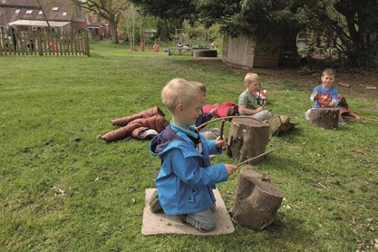 3 young children outdoors sitting on grass making music with sticks and bottom of tree trunk 