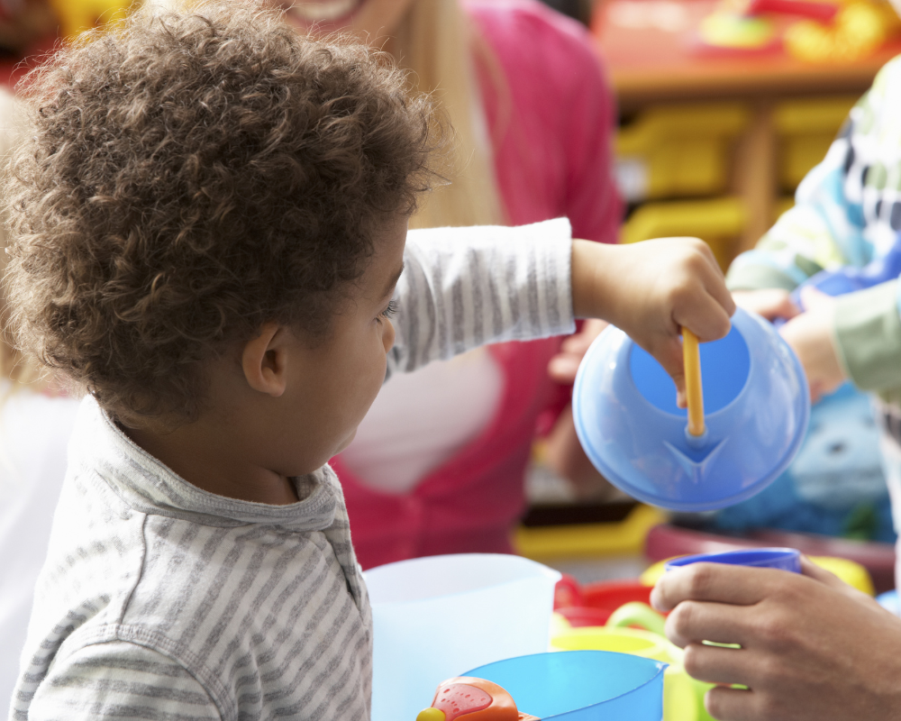 Young child playing in nursery school room with toy kettle, pretending to pour water in cup.