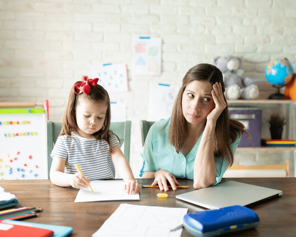 Stressed and tired woman sitting at the table next to young child drawing in a nursery setting