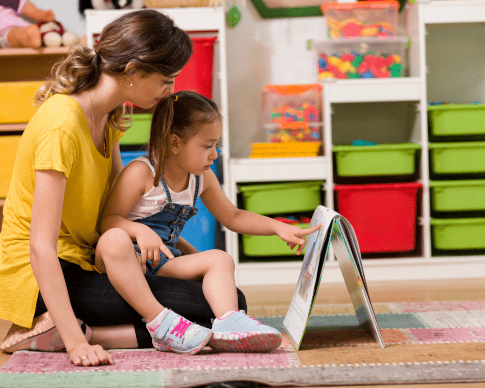 Little girl and teacher reading book together in nursery preschool classroom 