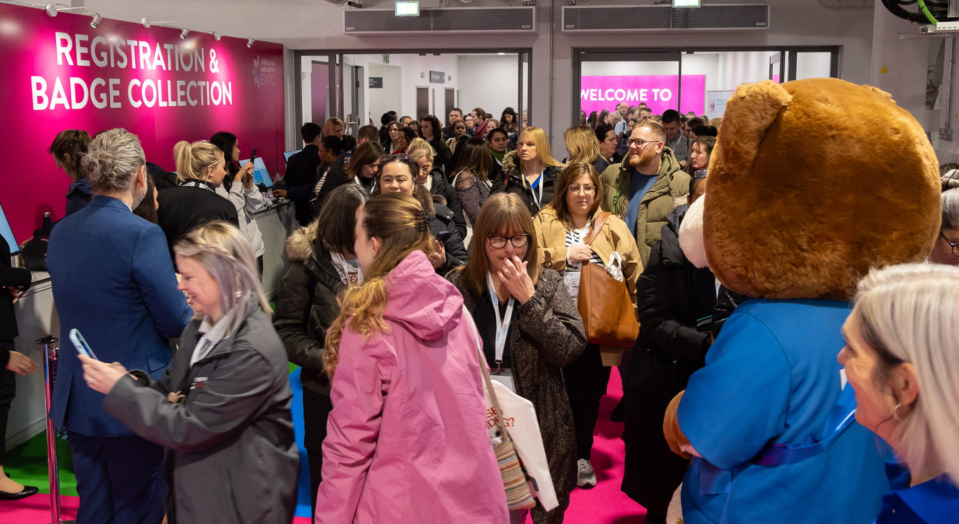 A cuddly teddy bear mascot wearing a blue t-shirt greets people coming into a childcare exhibition