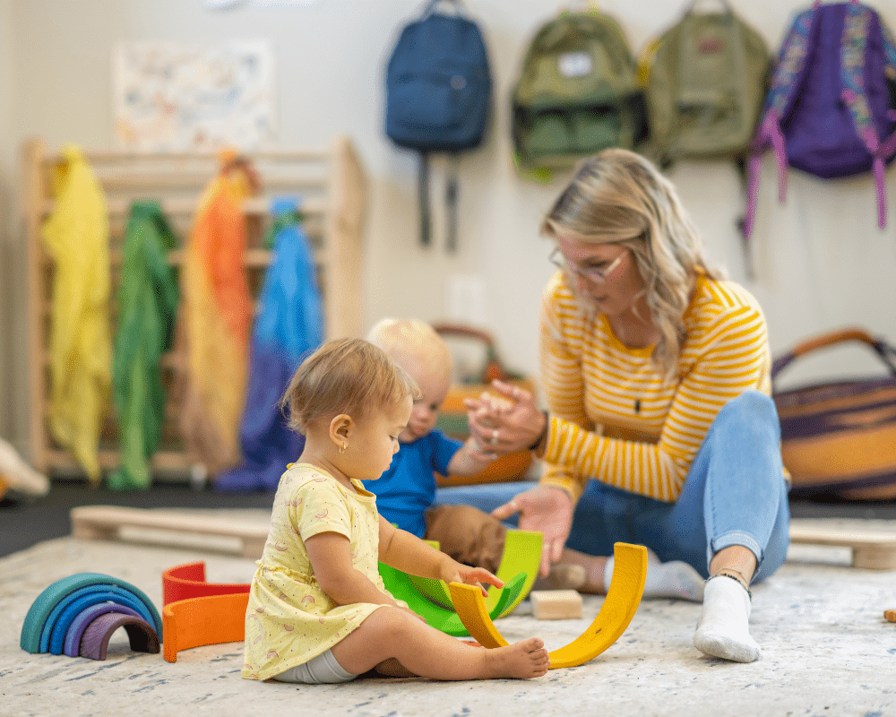 Two infants sit on the floor in their daycare room as they explore some colourful toys in front of them with their childcarer