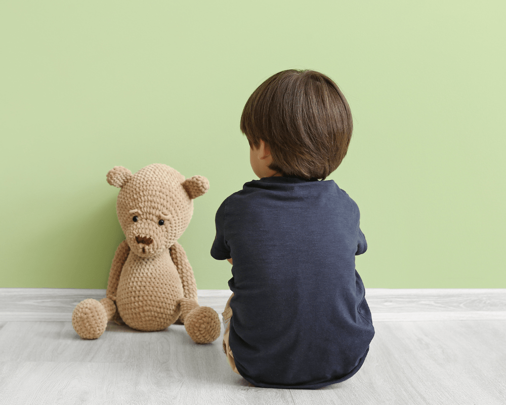 Little boy sitting near wall with back against the camera. A teddy bear with a sad expression sits next to him.