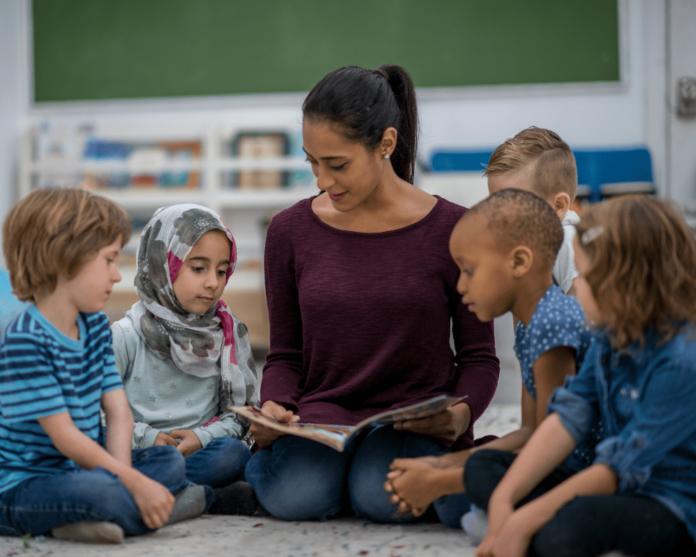 Multi-ethnic group of young children are indoors in their classroom listening to their teacher reading to them.