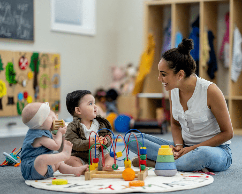 A small group of babies sit on the floor playing with various toys while their childcarer supervises and engages them