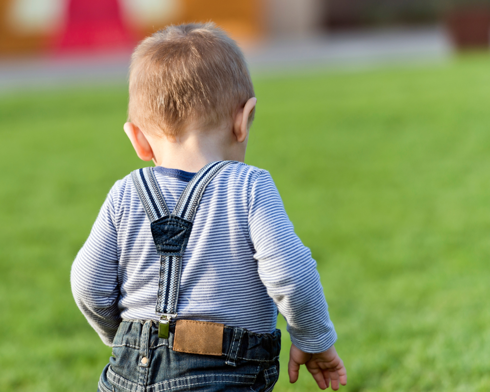 Toddler walking outdoors on the grass with his back to the camera 