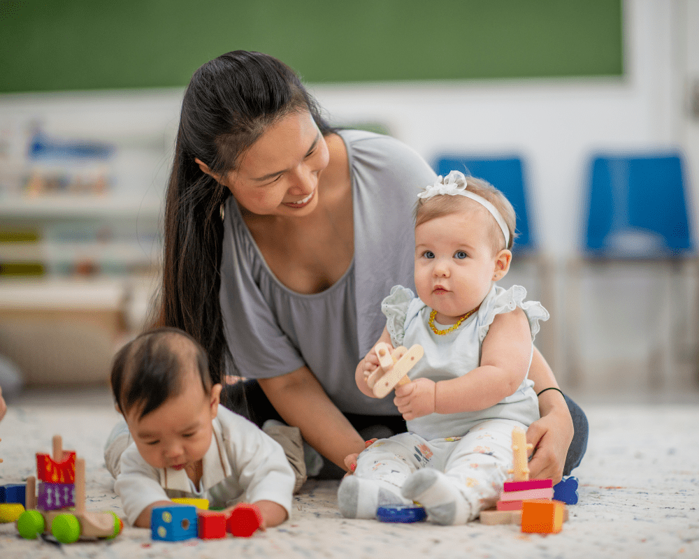 Childcare worker sitting on the floor, playing with two babies. The babies are playing with colourful blocks and toys.