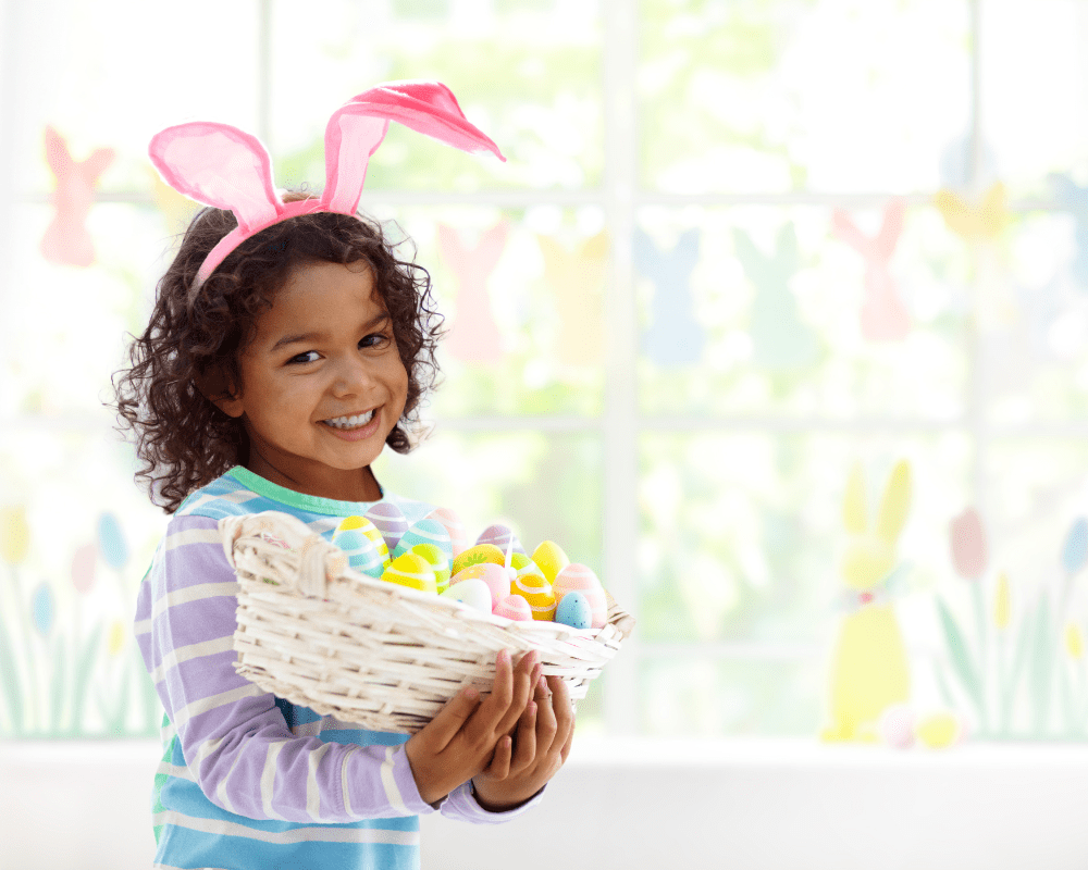 Young child wearing pink bunny ears whilst holding a white wicker basket with colourful Easter eggs inside