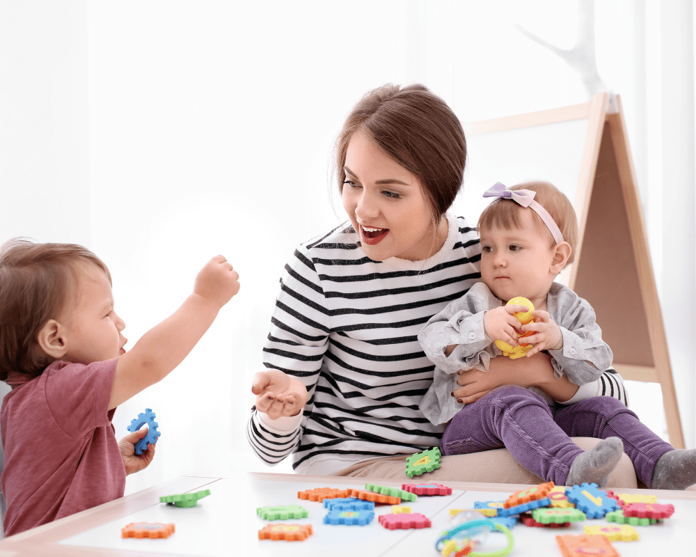 Childminder playing with two toddlers at table indoors, with colourful toys